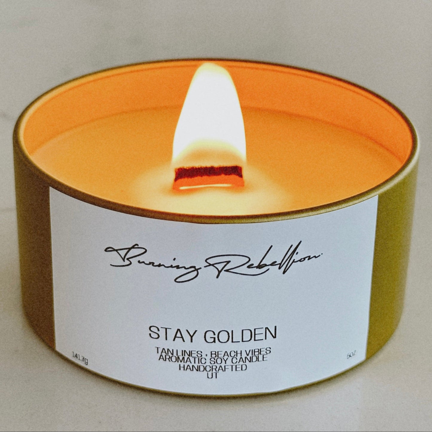 A lit Burning Rebellion candle labeled “Stay Golden” with a wooden wick in a gold tin, scented with tan lines and beach vibes.