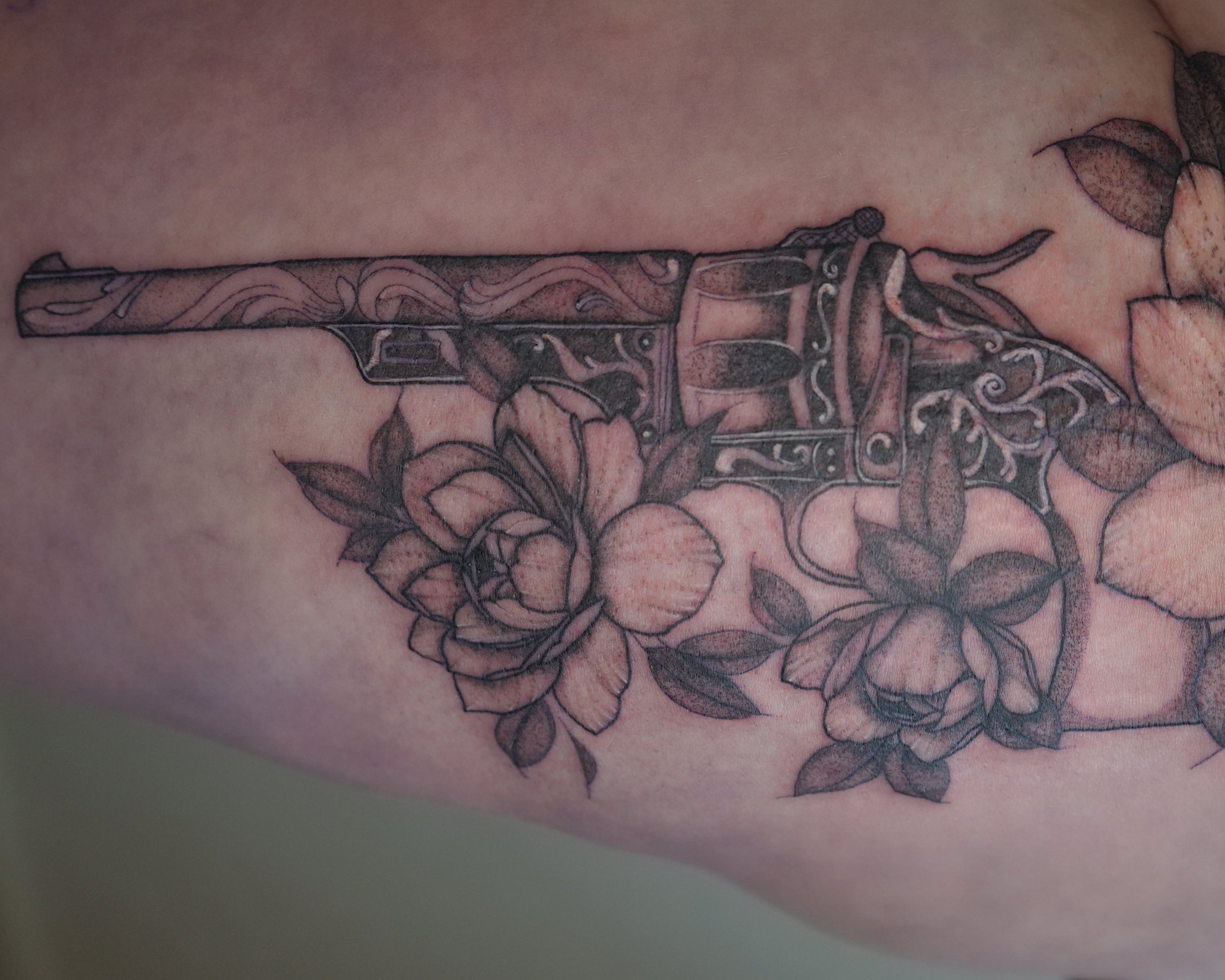 A close-up of a thigh featuring a black and gray tattoo design of a vintage key intertwined with large floral elements, shown on a person's leg.