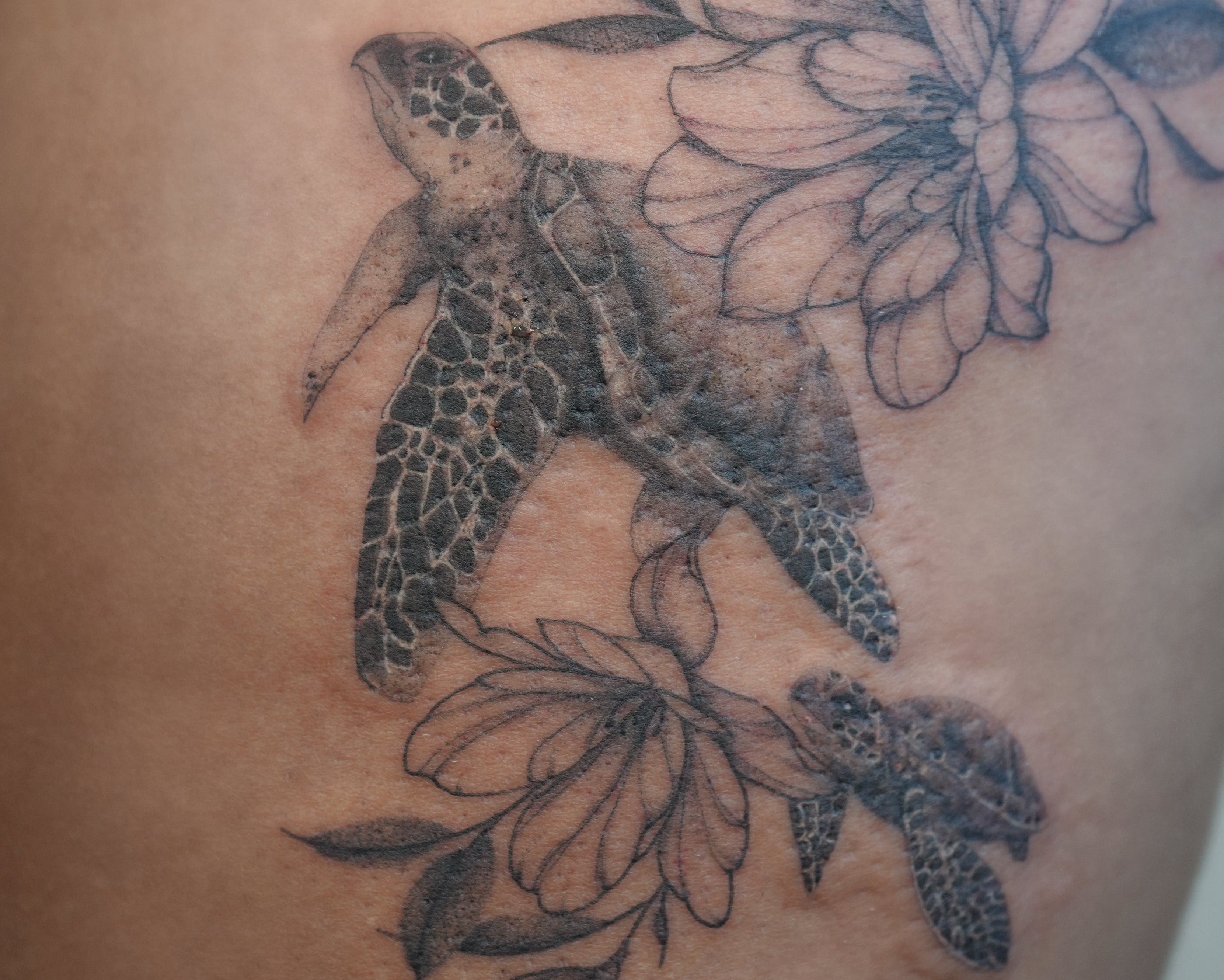 A black and gray tattoo of a sea turtle swimming among large flowers on a person's upper thigh, with a red garment visible at the top edge.