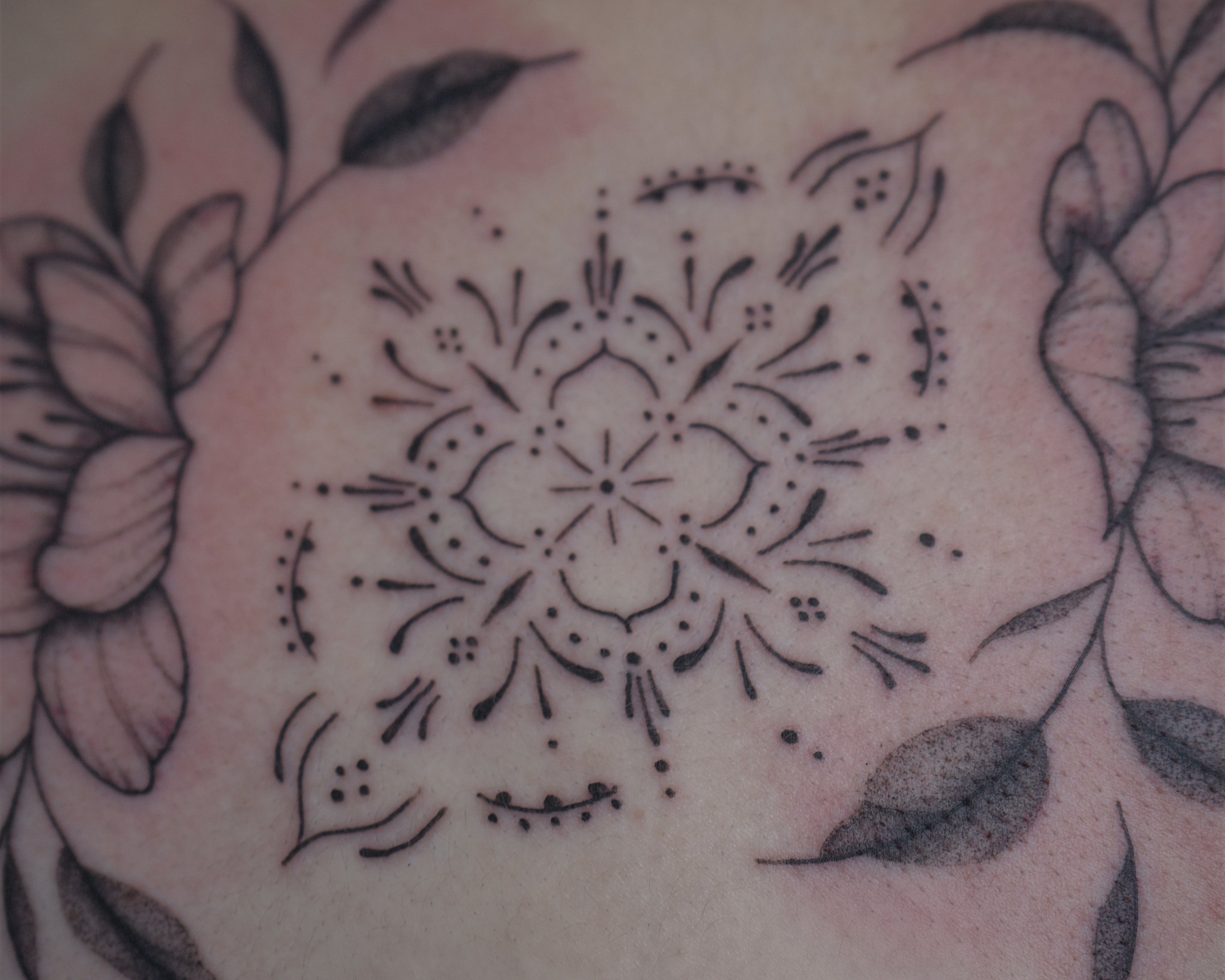 A close-up of a freshly tattooed skin area featuring a black mandala-style central design framed by floral line art on both sides.