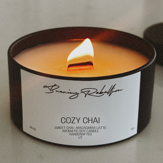 Candle in a black container labeled 'Cozy Chai' by Burning Rebellion on a neutral background.